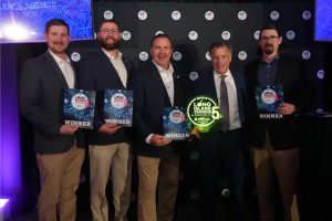Five men in suits pose on a stage, each holding a WINNER plaque at an awards event.