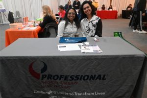 Two smiling women sit behind a booth table at a trade show, wearing white shirts, with brochures and a blue promo pouch on the table.