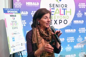 Woman speaking into a handheld microphone at a Senior Health & Beyond Expo backdrop with event banners.