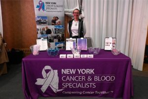 Booth for New York Cancer & Blood Specialists with a smiling representative behind a purple table, brochures and display materials on the table.