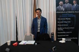 Man in a blue suit stands at a conference table with a banner advertising estate and life planning services beside him, smiling at the camera.