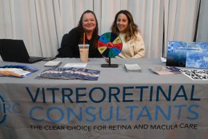Two women sit at a vendor table with a colorful spinning prize wheel, brochures, and a laptop, in front of a banner that reads 'VITREORETINAL CONSULTANTS' and 'THE CLEAR CHOICE FOR RETINA AND MACULA CARE'