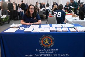A woman wearing glasses sits at a blue table with stacks of brochures at an information booth for Nassau County Office for the Aging during a crowded event.