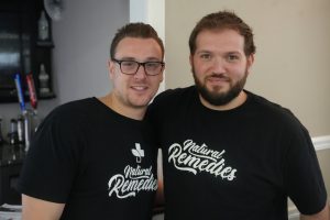 Two men in black shirts with 'Natural Remedies' logo, smiling at the camera in an indoor setting.