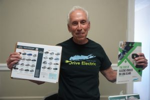 Smiling man with white hair in a black Drive Electric Long Island shirt, holding an electric-vehicle guide in one hand and a green-and-white EV pamphlet in the other.
