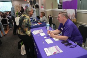 Older woman speaks with a health fair volunteer at a booth with purple tablecloth and brochures nearby.