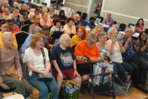 Largest group of seniors seated in rows at an indoor community event, many smiling and taking photos with phones.