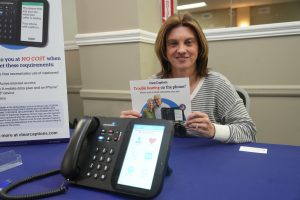 Smiling woman sits at a table holding a brochure that reads 'Trouble hearing on the phone?' beside a large conference phone on a blue table surface.