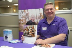 Smiling man in a purple polo sits at a table displaying Aetna materials and brochures with a purple banner behind him.