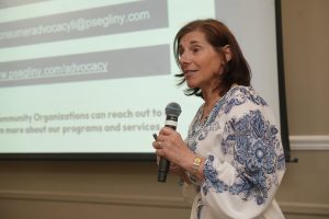 Woman speaking into a handheld microphone during a presentation, standing beside a projected slide with blurred text in a conference room, wearing a blue and white patterned blouse and a gold watch.