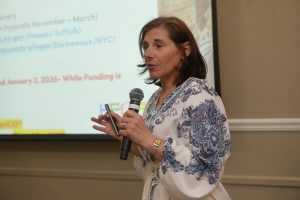 Woman in a white and blue patterned blouse speaks into a handheld microphone during a presentation, with slides in the background.