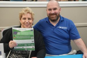Smiling woman holding a green flyer about home energy programs, seated next to a man in a blue Department of Public Service shirt.