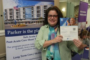 Smiling woman with red glasses holds a Parker Jewish Institute brochure beside a large campus poster in a booth.