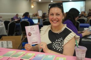 Smiling woman at a conference booth holding a pink Vollmer Senior Consulting LLC flyer, with colorful notepads on a pink table in front.