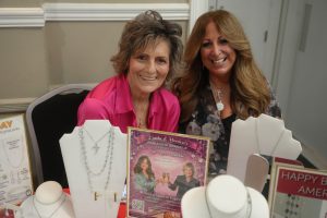 Two smiling women at a jewelry booth; foreground shows necklace displays and promotional flyers visually behind them.