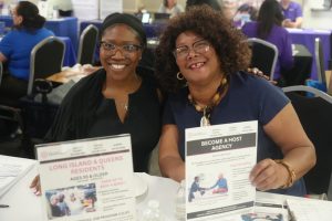 Two smiling women sit at a table with brochures at a busy community event.