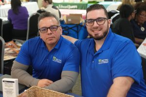Two men in matching blue polo shirts sit at a booth table, smiling at the camera at a conference event.