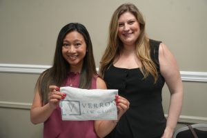 Two women smiling indoors, holding a light gray cloth with a law firm logo between them.
