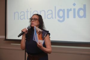 Woman speaking into a handheld microphone, holding brochures, in a room with a large blurred 'national guard' projection behind her.