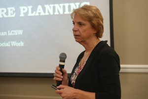 Professional woman in a black blazer speaks into a handheld microphone during a presentation, with a projected slide behind her.