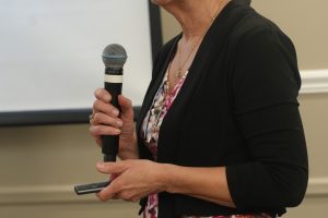 Woman in a black blazer and floral top speaks into a handheld microphone during a presentation.