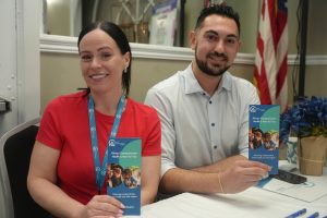 Two smiling volunteers sit at a table with blue pamphlets about compassionate health, woman in red shirt and man in light shirt, US flag in background.