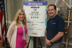 Two adults pose beside a large sponsor board for a health expo, with an American flag in the background and logos listed on the board.