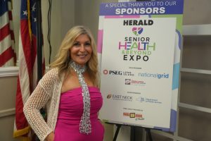 Smiling woman in a pink dress with a silver scarf stands beside a sponsor board at a health expo.