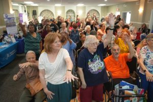 Large group of seniors in a bright hall raising hands as they participate in a community event.