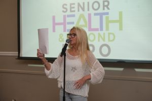 Woman with blonde hair, wearing a white knit sweater and glasses, speaks into a microphone while holding a sheet of paper at a presentation.