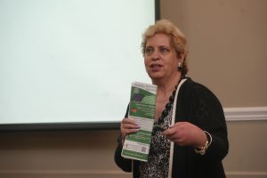 Woman presenter in a black cardigan holds a green energy brochure while speaking to an audience.