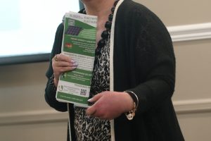 Female presenter in a black cardigan speaks at a seminar, holding a green energy brochure.