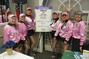 Six women in pink satin jackets and pink caps pose beside a sponsor board at the Senior Health & Beyond Expo.