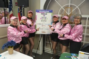 Six women in pink jackets and hats pose around a large sponsor board for the Senior Health & Beyond Expo.