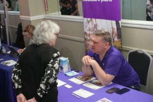 Older man in a purple polo talks with an elderly woman with white hair at a booth; brochures lie on a purple tablecloth behind them.