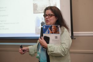 Woman wearing glasses speaks into a handheld microphone while presenting, holding a brochure and a smartphone in front of a projected slide.