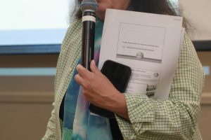 Woman speaking into a handheld microphone while holding printed handouts and a smartphone.