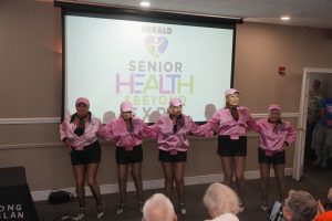 Six senior women in pink jackets and hats pose in a line-dance formation on a stage, with a projection reading 'HERALD Senior Health & Beyond Expo' behind them.