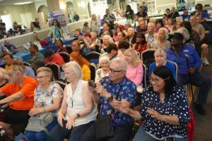Large crowd of mostly elderly people seated in a conference hall for a community event, with booths and posters in the background.