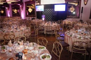 Banquet hall set for a formal event with round tables, gold chairs, beige tablecloths, candle centerpieces, and a stage with two projection screens in the background.