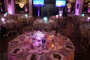 Round banquet tables set for an awards dinner in a dimly lit ballroom, with pink-purple uplighting, gold chairs, and a stage with podium and screens.