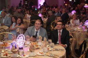 Two men in suits sit at a round banquet table in a crowded formal event, with pink neon centerpieces and plates of food nearby.