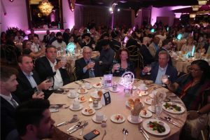 Group of diverse guests at a round table toasting with glasses during a formal gala dinner, with pink lighting and elegant place settings in the background.
