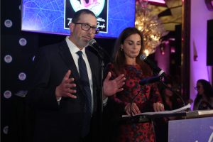 Man in a dark suit and glasses speaks into a microphone at a podium, while a woman in a red floral dress stands beside him with notes on the lectern.