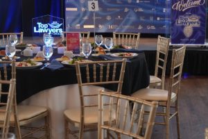Banquet hall set for a 'Top Lawyers' awards event with round tables, gold chairs, place settings, and a blue backdrop featuring the logo.
