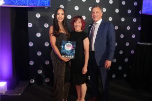 Three adults posing for a photo at an awards event; the woman on the left in a glittery black jumpsuit holds a 'WINNER' plaque, flanked by a woman in a black dress and a man in a blue suit; backdrop features circular logos.