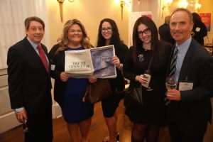 Group of five adults in business attire posing for a photo at a formal event, one holding a printed flier that says 'TAKE THE CHAOS OUT OF DEPOSITIONS' and another with a poster, drinks in hand in a warmly lit room.