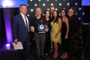 Eight people pose on a stage at an awards event; the man in a black polo holds a 'WINNER' plaque.