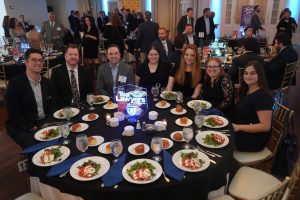 Eight professionals in formal attire sit around a round banquet table with salad, bread rolls, and water glasses; a glowing centerpiece reads 'Top Lawyers'.