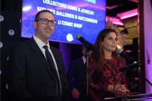 Two presenters at a podium with microphones on a stage, a man in a suit and a woman in a floral dress, with purple lighting in the background.